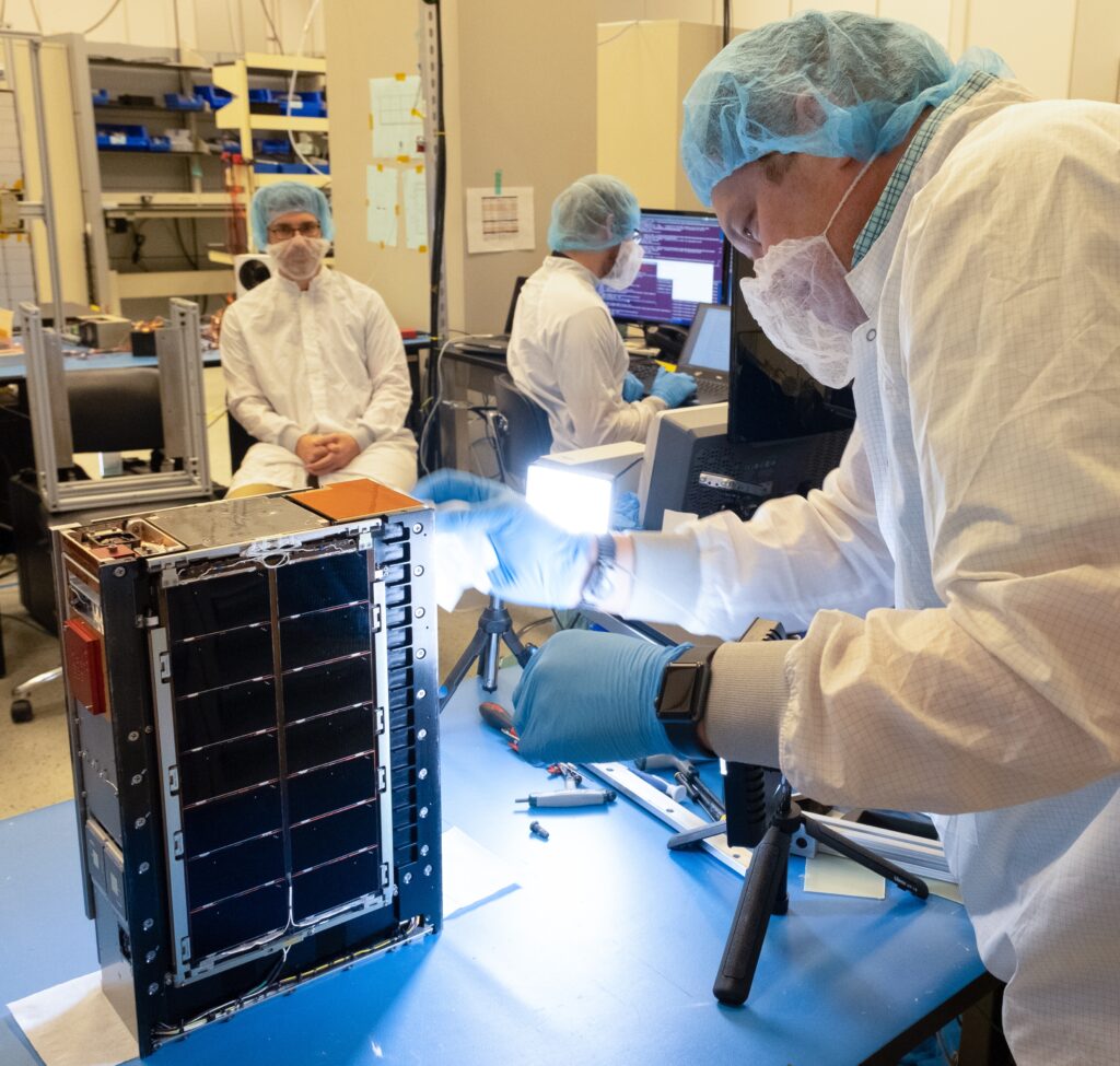 Several technicians in white smocks, masks and head coverings work around a small briefcase-sized piece of hardware.