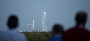 A large, white rocket stands vertical on a launch pad, a thin trail of white exhaust exiting toward the middle of the rocket. A tall, white water tower stands next to the launch pad against a pale blue sky. In the foreground, blurred out, are the heads of shoulders of people looking at the rocket.