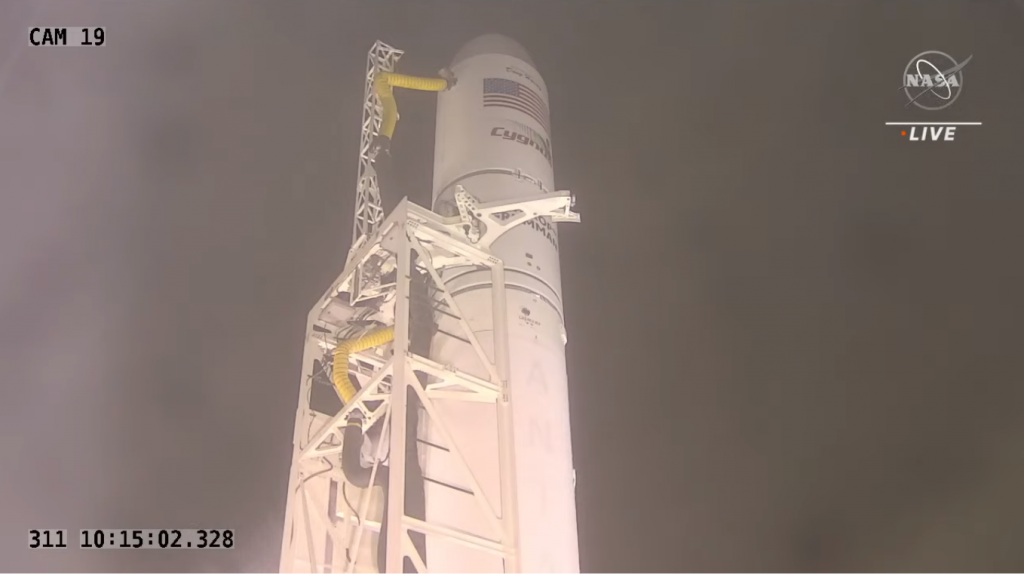 against a dark pre-dawn sky: white Antares rocket, with American flag and "Antares" printed near the nose; rocket is supported by white-hued scaffolding