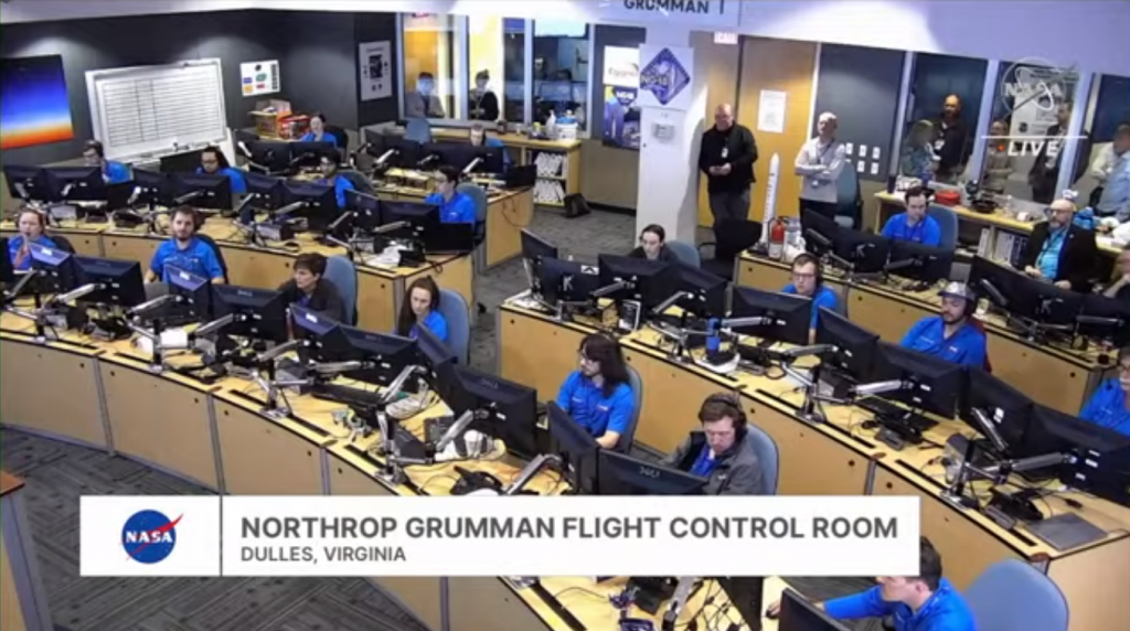 wide view of a control room, with people mostly clad in blue sitting at light brown desks