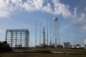 A large, white rocket with the words “ANTARES” in black letters stands vertical on its launch pad, surrounded by four poles used for lightning protection. To the right of the rocket, a water tower with long, spindly white legs has a white dome on top, with a dark logo that’s not fully visible. To the left of the rocket, is a large, open metal structure with a smaller circular structure on its side. There are several tall, gray light poles placed around the road. All of this is framed against a light blue sky with fluffy white clouds.