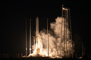 A photo of a rocket launching at night. The Antares rocket is slim and white with a short, rounded nose cone. It rises off the pad with brightly lit white clouds billowing around it and beside it against a black sky. A tall water tower is just visible in front of the clouds to the right of the image.