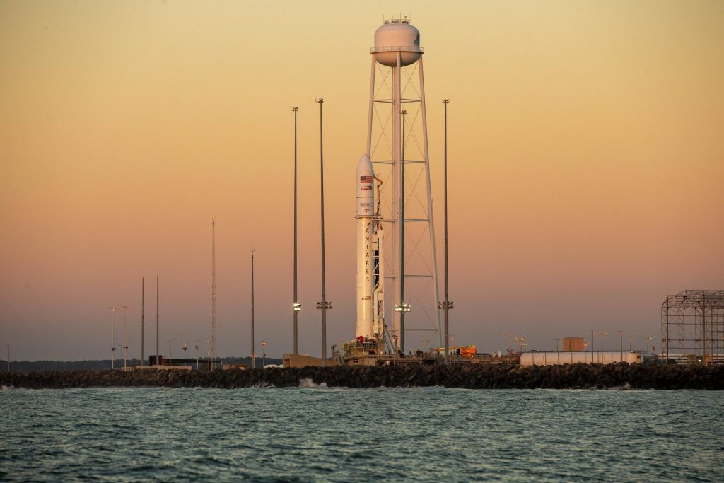 golden sky backdrop with white Antares rocket in center of frame, along with water tower and lightning suppression towers