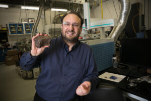 A white person with short, brown hair, wire-rimmed glasses, a smile, and a beard stands in the foreground of this image. This is Jason Dworkin, an astrobiologist at NASA's Goddard Space Flight Center in Greenbelt, Maryland. Dworkin is the project scientist for NASA's OSIRIS-RE mission. He is visible from the waist up, wearing a dark blue dress shirt and holding up a brown rock with his right hand. The rock is a fragment of the Canyon Diablo meteorite, which struck Earth 50,000 years ago, creating Meteor Crater in Arizona. In the background of the image is Dworkin's lab at NASA Goddard, featuring blocky, metal machines, plus wires and metal tubes handing from the ceiling.