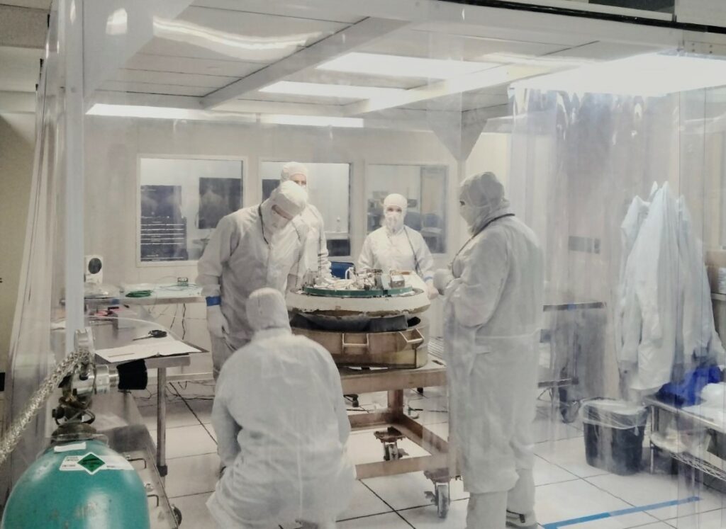 The image shows five scientists covered head to toe in white suits, with long robes, and face coverings. One person has his back to the viewer, and is crouching in the left bottom corner. The rest are standing in a circle around a large metallic object, a mock sample capsule. The capsule is on a cart in the middle of the room. Inside this cleanroom everything is white, including the neon lights on the ceiling. The scientists are practicing opening a capsule with asteroid samples in it. The real capsule with samples from asteroid Bennu, is on its way to Earth now.