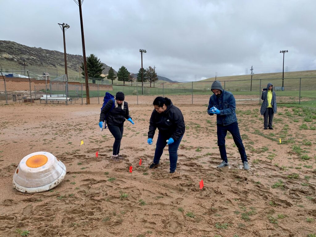 Four people are standing in a muddy field outside of Denver, Colo., surrounded by metal fencing. They're dressed for cold weather and all wearing nylon gloves. They're all focused on an object in the bottom left corner of the image. The object is shaped like a lampshade -- it's a mock sample capsule. One of the team members is standing by themselves in the right hand corner of the image, holding a video camera that's pointed at the rest of the team and capsule.