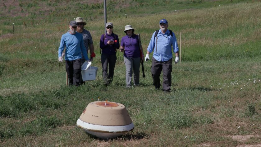 Five people approach a brown and white capsule sitting on a grassy field.