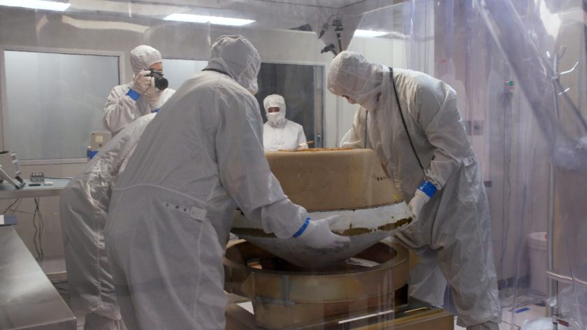 People clothed in white from head-to-toe hold a brown capsule in a clean room.