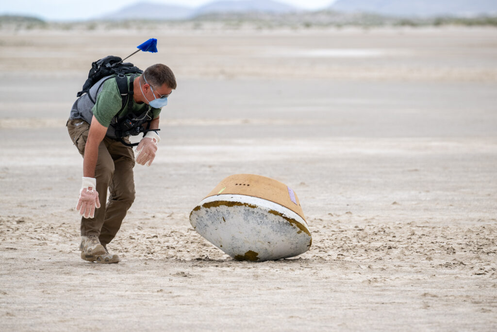 On a beige expanse, a person is pictured wearing a backpack, gloves, and face mask. He is crouching over a mini-fridge size object shaped like a badminton birdie.