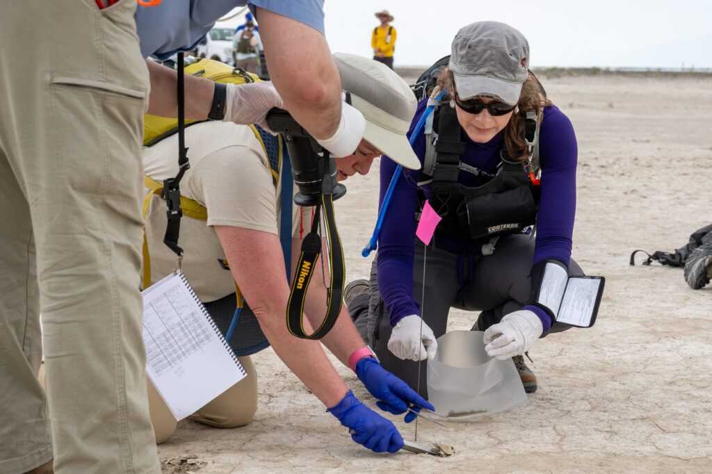 Two people, wearing caps, sunglasses, and latex gloves, are bent on their knees over a sandy surface. One is holding a small, plastic baggie, while the other is scooping some sand. Another person, with only an elbow and part of the right leg visible, hovers over them in the left-hand side of the image, taking a photo.