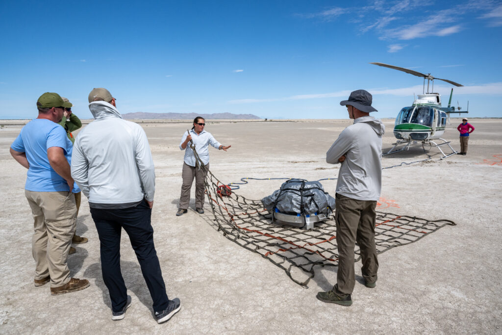 In the foreground several people are standing with their backs to us. In the background a person is facing us, holding the corner of a large piece of mesh, with a tarp-covered object on top of it. The white ground makes for a beautiful contrast with the bright blue sky. In the right-hand corner stands a helicopter, and next to it, a person with their hands on their hips is facing us.