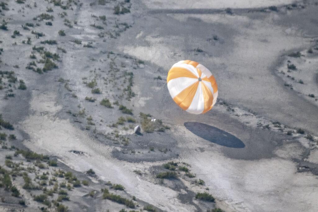 A round, striped parachute is pictured drifting toward the desert ground. Attached to the parachute is a long cable, which is carrying a cone-shaped capsule at the bottom. 