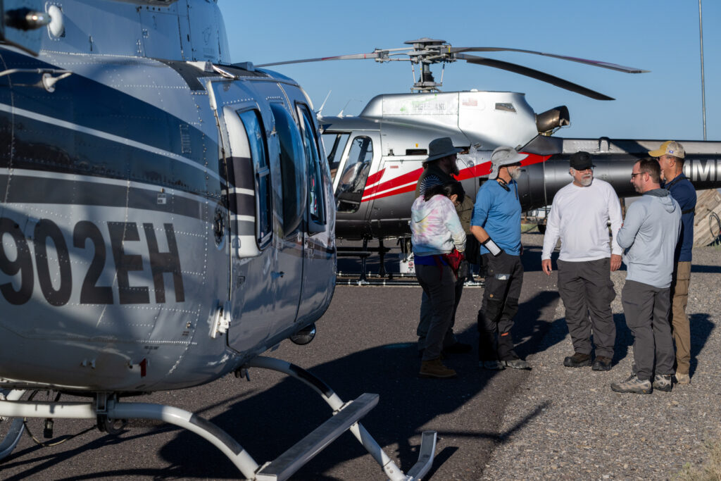 A helicopter is shown in the left forefront. Several figures huddle in the far right. Behind them is another helicopter.