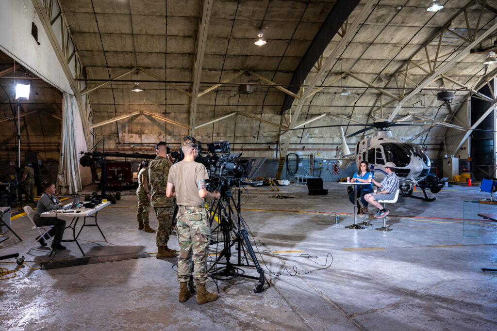 Several figures stand in a line on a cement floor in the forefront of the image. They are standing behind video cameras mounted on tripods. A tall, domed ceiling is visible high above them. About 10 feet in front them sit two figures on stools behind a tall table. Behind the people sitting at the table is a helicopter.