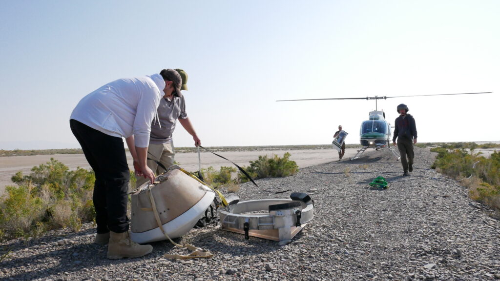 In the forefront, on a rugged strop of land, two figures are handling a cone-shaped object. In the background is a bright, day sky, and beneath it a sandy desert. Two figures are seen walking toward the viewer, with a helicopter behind them.