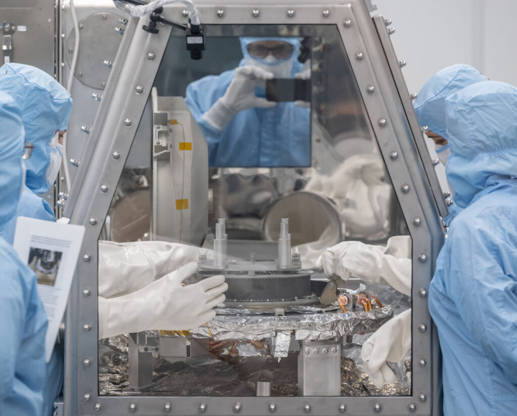 Five people in blue clean room gowns and white gloves stand around a glove box with a circular sample head inside.