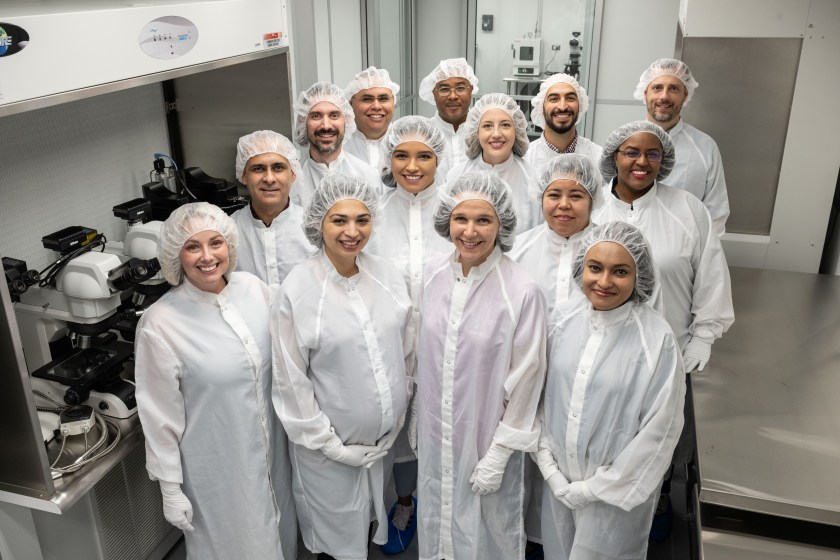 A group of thirteen men and women in a lab dressed in white clean room gowns, white gloves, and white hair coverings. 