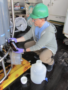 A scientist is crouching in front of a gray tank holding a small brown glass bottle. The scientist is wearing purple latex gloves and a green hard hat to safely perform the research aboard the ship.