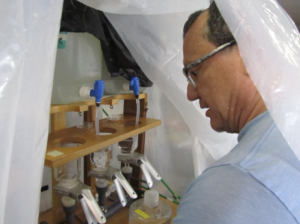 A scientist is standing under a plastic sheet that serves as a barrier for his experiment station in front of him. The experiment includes several containers of liquid and plastic tubing.