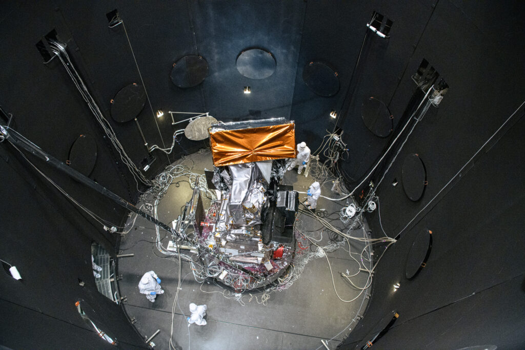 The perspective of the image is from the top of the chamber peering down, looking into the chamber at the observatory. The inner walls of the test chamber are black, and the observatory, centered in the image and in the chamber, contrasts that darkness with copper-colored reflective material shining against the black. There are four scientists in white protective clean suits around the observatory and wires scattered on the floor connecting to the observatory and the chamber walls.