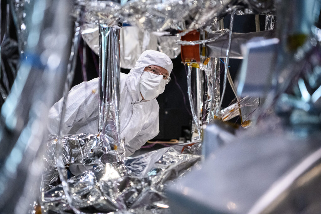 The camera peers through parts of the observatory, focusing on an engineer who is looking intently at parts of the observatory. The engineer is wearing white protective clean gear that covers up over his head, and also includes a mask, so the only part of his face that is seen are his glasses-covered eyes. The parts of the observatory surrounding the scientist are a glimmering silver color.