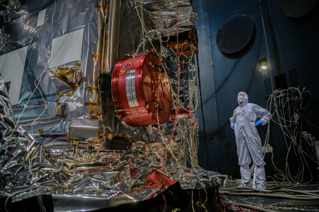 A majority of the image is taken up by the observatory, primarily from the left side of the image. The complex pieces of the observatory are surrounded by wires and reflective foil-like material. Centered in the image is a red piece of equipment on the observatory, stoutly cylindrically shaped. To the right of the image stands a scientist in a full white protective suit. They stand feet slightly apart with hands resting on their hips, in a superman-like pose. 