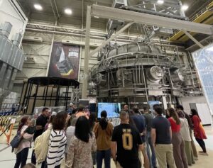 A large group of people stand in the foreground of the image, facing away from the camera. They are facing a large metal chamber - the thermal vacuum chamber - which has several grey, metal tubes on the side. The chamber takes up the full top right corner of the image.