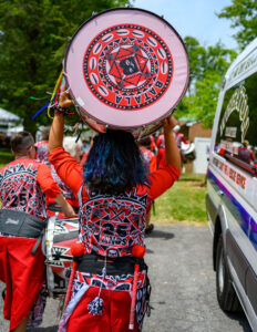 A woman is centered in the image wearing a bright red outfit with white, black, and blue patterns and designs in circular shape on the back of her outfit. The woman has down black hair with blue streaks through it. She is carrying a drum that rests on the top of her head. The drum has the same pattern of red, black, white, and blue colors on its face.