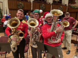 Three men stand in the picture. All are holding euphoniums in their hands. From left to right, Gary is the third man in the picture. He is wearing a red sweater and has a red Santa hat on. The man in the middle also wears a red sweater and has a green holiday had on. The man to the left is wearing a military outfit with a red jacket and black pants. They are standing in a large auditorium and wreaths can be seen hanging in the background.