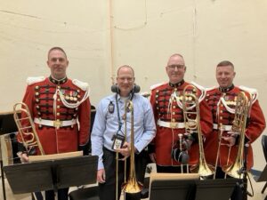 Four men stand in a picture. All four are holding trombones in their hands. From left to right, the second man is Gary Davis. He wears a light blue long-sleeved button up shirt and has protective headphones around his neck. The men he is standing next to are all wearing military uniforms with red jackets, white belts, and black pants. 