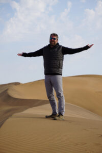 A man stands centered in the image standing straight with his arms held out on either side of him. He is standing on a tan sand dune. The sky behind him is a light blue and gray color.