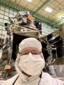 A man, seen from his shoulders up, takes up a majority of the picture. He is wearing a white clean suit which covers over his head, and also a white mask that covers his nose and mouth. He has glasses on. Behind him is the PACE spacecraft which is very large and covered in wires and other metallic materials. 