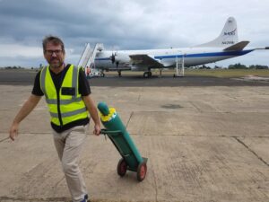 A man takes up the left side of the image. He is walking towards the camera. He wears a neon yellow vest and is holding onto a large, green gas canister, which is rolling behind him. In the background of the image, a large plane sits on a runway with the nose of the plane facing the left of the image and the tail of the plane, featuring a NASA logo, on the right side. The plane has a stairway connected to the door.