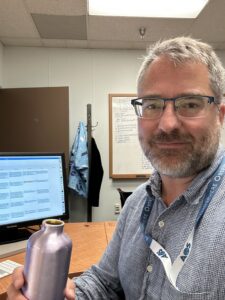 A man is sitting at a desk in an office. He is facing the camera and appears to be taking a selfie. He takes up the right side of the image. The left side of the image shows a computer screen and a water bottle, which the man is holding. In the background, the office door, a coat rack, and part of a whiteboard can be seen.