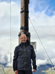 A man stands centered in the image and can be seen from the waist up. He is wearing a black winter coat. Behind him, also centered in the image, is a large wooden pole. There are wires coming off of the pole. In the background of the image, the sky is bright blue, but is covered by some white fluffy clouds. Near the bottom of the image in the background, a mountainous green landscape can be seen far below where the man is standing.