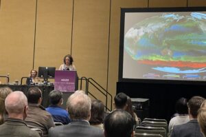 A woman in a light-colored blazer stands behind a podium on a dais, giving a presentation. On the right side of the image, a screen shows a colorful visualization of Earth, with concentrations of different types of phytoplankton in the oceans represented with different colors. In the foreground of the image are the backs of attendees’ heads.