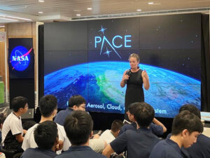 A woman stands in front of a set of nine screens, which together show an image of part of the Earth, with the PACE logo above it. She is wearing a black dress and holds a microphone. The woman is facing a group of people who have their backs to the camera and are watching the woman present.