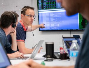 The image is focused in on a man to the left of the image wearing glasses and a blue and red short sleeved polo short. He wears a headset, where the wire is hanging down past the far side of his head. He is pointing at a computer screen which is in the background of the image. In front of him, but out of focus in the iamge are two other people, one sitting next to the man and one sitting across from him, closer to the camera. At the table they are sitting at are computers, coffee mugs, and water bottles. 