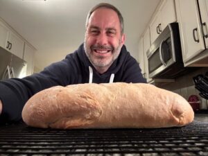 A man wearing a black hoodie takes a selfie. In front of him is a loaf of light tan, freshly baked bread, which takes up the entire length of the image, left to right. The bread is resting on a cooling rack.