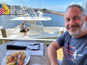 A man wearing a blue T-shirt with a spaceship printed on it, takes a selfie, taking up the right side of the image. He is sitting at a table at a restaurant with a plate of food in front of him. In the background of the image is a white boat with one man sitting on it. It is floating in a harbor, the water calm and blue.