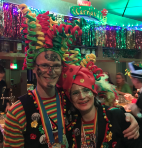 A man and a woman are standing side by side, the man to the left with his arm around the woman's shoulder. They both have glasses on and their faces are covered in a glittery face paint. They wear green, yellow, and red striped shirts and black vests that have colorful puns on them. They both have large hats upon their heads with green, yellow, and red spirals on them. The lighting in the room is dim and there are streamers hanging on the walls behind them. 