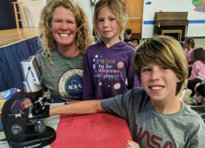 A woman and two children are seen in the picture. The woman, with blonde curly hair, is to the left of the image, wearing a gray shirt with a NASA meatball logo on it. In the middle is a young girl wearing a purple shirt that says, "I want to be a princess and a physicist" with planets surrounding the words. To the right is a young boy with blond hair who wears a gray T-shirt with the NASA worm logo on it. He is reaching forward and holding a microscope.