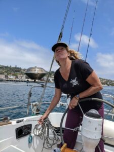 A woman stands slightly to the right of center of the image. She is wearing a black T-shirt and has a hat on. She looks to the left of the image, out in the distance. She is standing on a boat and is holding the steering wheel in her left hand and rope in her right hand. Behind her is the ocean, part of a harbor, and a blue sky with some clouds.