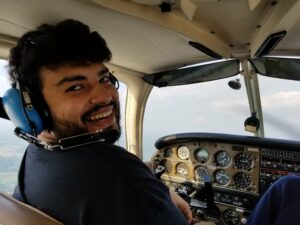 A man wearing a dark shirt and a headset with a microphone attached in front of his mouth sits in the cockpit of a small airplane. He is seen from the shoulders up. The image is being taken from behind him, so he is turned around, smiling at the camera. In front of him are the controls of the plane including six circular gauges. Out of the front window of the plane is a gray sky.