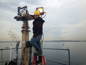 A man stands at the top of a small orange step ladder, centered in the image. He wears jeans, a dark colored jacket, and a yellow hard hat. He is standing next to a structure that is about the same height as him and has a scientific instrument on top of it. In the background of the image is a body of water, which is blue and still. The sky is cloudy and is a light blue and gray color.