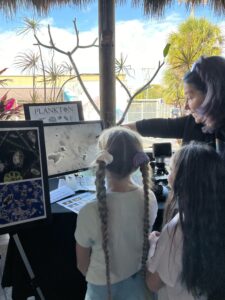 Two children stand in the foreground of the image facing away from the camera. They are looking at a screen in front of them. To their right is a woman with blue and purple hair pointing to the screen. On the screen are several blurry blobs - phytoplankton - which are being displayed from the microscope, seen between the heads of the two children. 