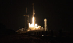 Centered in the image is a rocket, mid-launch. The launch is taking place at night, so all around the rocket and it's stand is black. The only light is coming from the fire emerging from the bottom of the rocket in an orange glowing color, illuminating a cloud of smoke coming from the rocket as well.