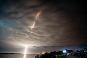 The image primarily is focused on the sky, which is dark and cloudy at night. There is a streak of light that starts near the bottom left corner and rises up to the center top of the image. The clouds surrounding this streak glow a brighter white than those that aren't illuminated by the light. The light is also reflected off of the ocean, which is seen in a small strip at the bottom of the image. 