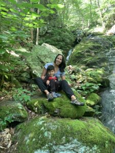 A woman with dark hair sits on a moss covered rock with a small boy sitting between her legs. They are centered in the image and smiling at the camera. Behind them are several other moss covered boulders and lots of leaves and foliage. Much of the image is a bright green color because of the greenery.