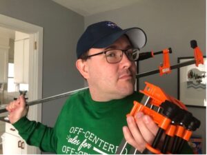 A man stands centered in the image wearing glasses, a green crewneck sweatshirt, and a baseball cap. He holds four woodworking tools - clamps - in his left hand. In his right hand he has another clamp which is resting on his shoulder and extends behind his head. The clamps are orange, black, and silver colored in their pieces.